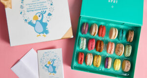 A box full of assorted French macarons has a French macaron box with a custom sleeve and a customized greeting card to its left.