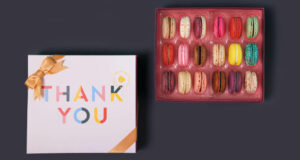 A box full of assorted French macarons has a French macaron box with a Thank You sleeve to its left.