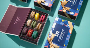 A box full of 9 assorted French macarons is surrounded by French macaron boxes with custom sleeves.